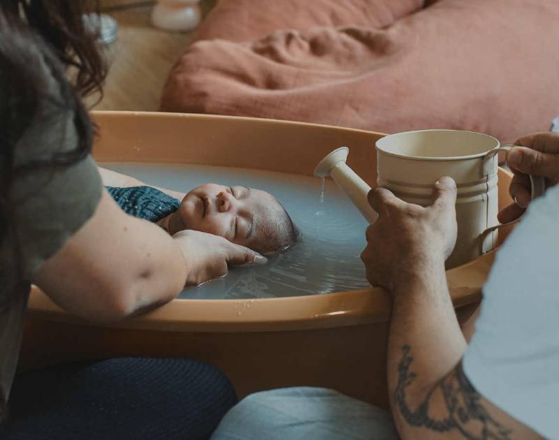 visuel signature, bébé plongé dans une baignoire, sa mère le tient, un homme verse un filet d'eau sur son crane